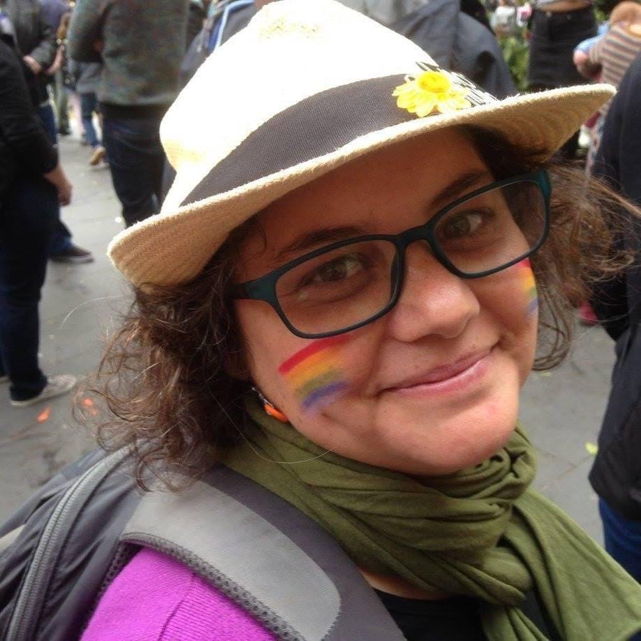 This image is of a women’s head. She is looking into the camera; she is wearing white hat with a black band. She has curly hair around the sides of her face. She is wearing glasses and has a rainbow painted on her face. There are bodies in the background, indicating that she is in a crowd.