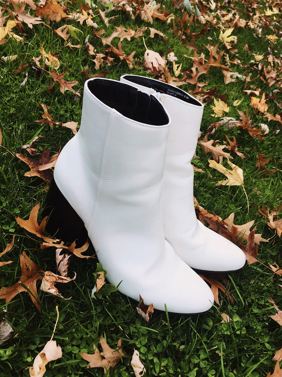 White ankle boots with a three inch heel placed on the grass surrounded by fall leaves.