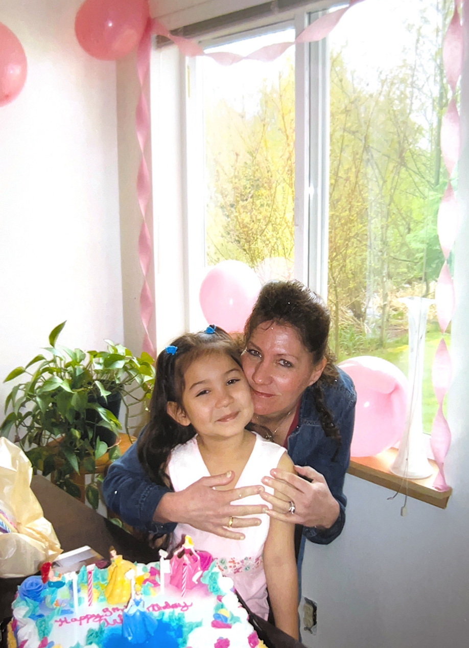 A young Aria and her grandmother post for a photo at Aria’s birthday party. The pair sit in front of a large birthday cake surrounded by pink balloons and streamers.