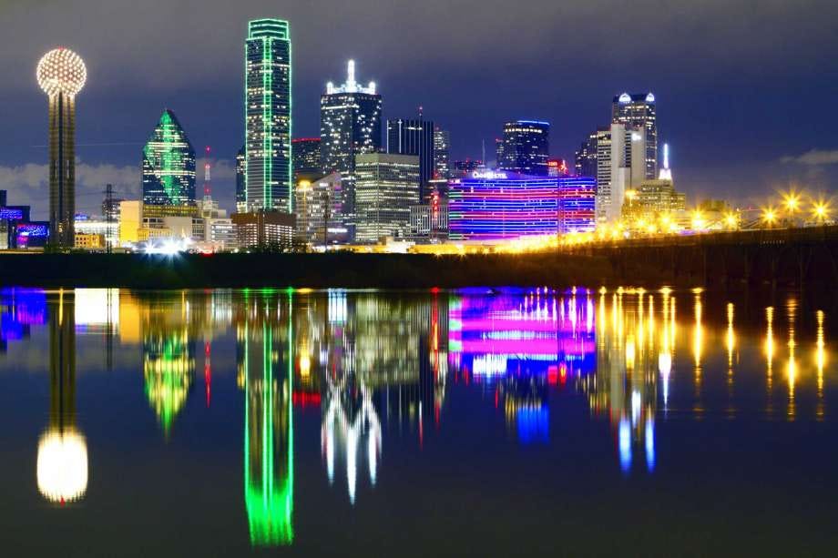 Dallas city skyline. Various buildings are lit in different colors, with a walkway bridge lit with globe lights, over water.