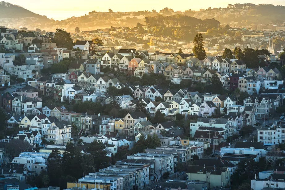 Rows of colorful homes, mostly in shade, are interspersed with trees in San Francisco. Muted sunlight filters across them.