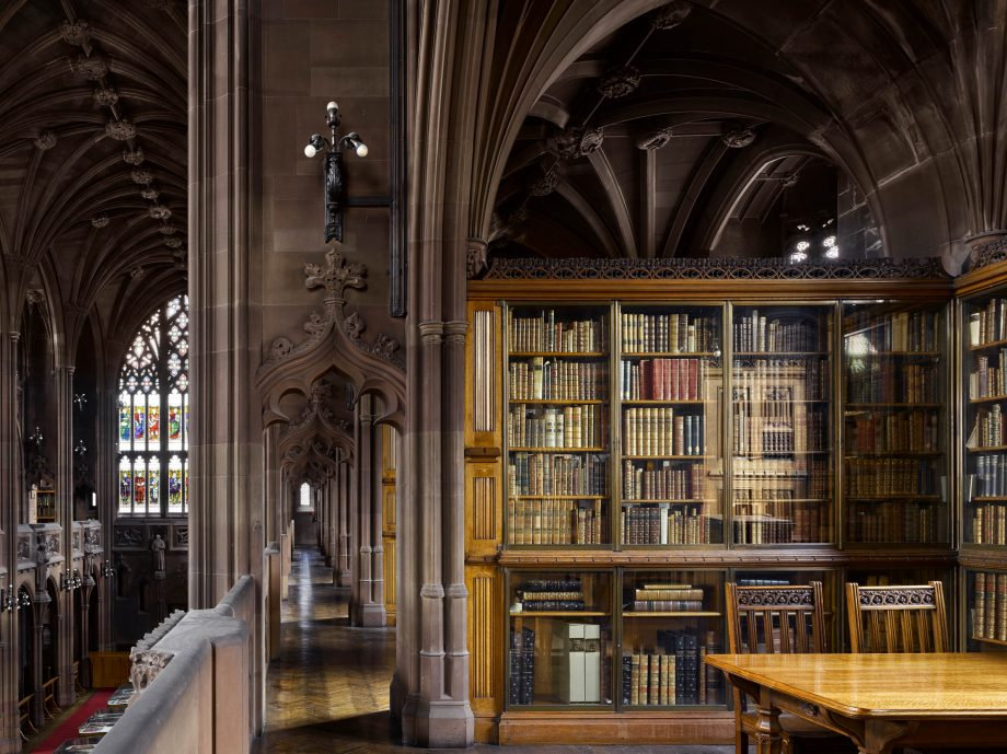 A neo-Gothic style library. Upstairs with a hallway and arched walls and pillars. On the right side, a book-case of rare books and a wooden table on the bottom right with two chairs.