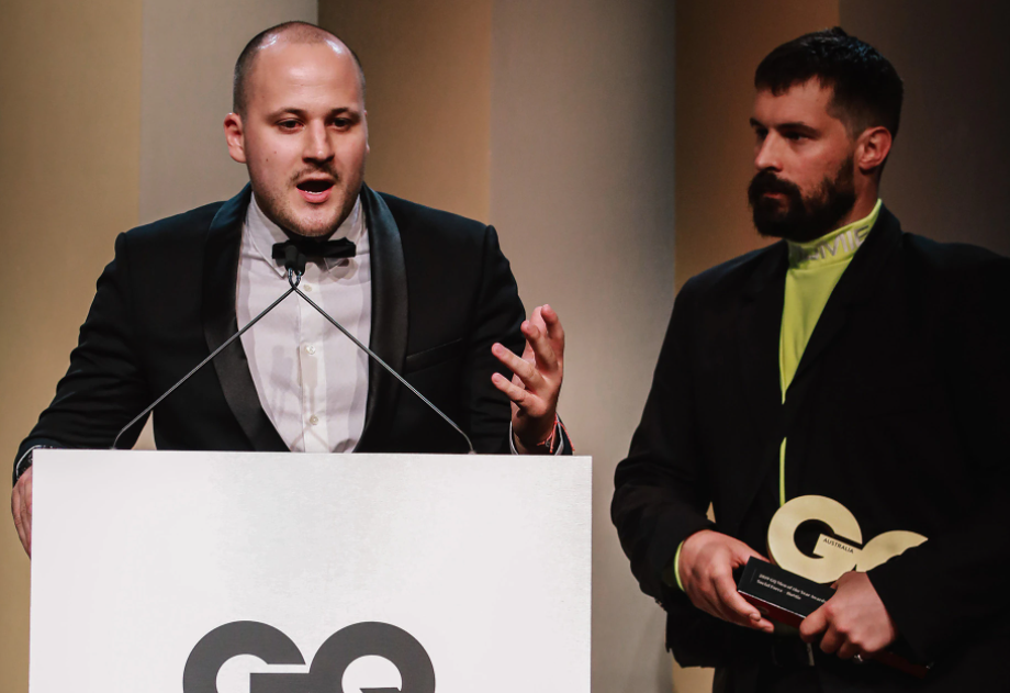 Nick Pearce and Marcus Cook stand at a podium while they accept their GQ award. Nick is wearing a tuxedo and speaking into a microphone. Marcus is standing next to him, holding their award.