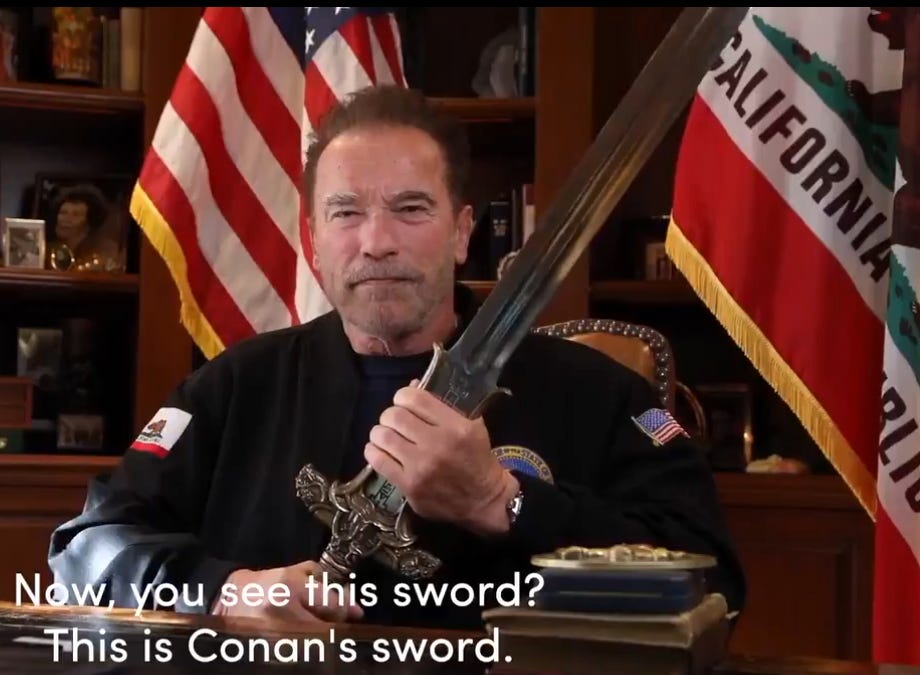 Seated Arnold Schwarzenegger gripping movie prop sword, wearing dark blue shirt emblazoned with US and California flags.