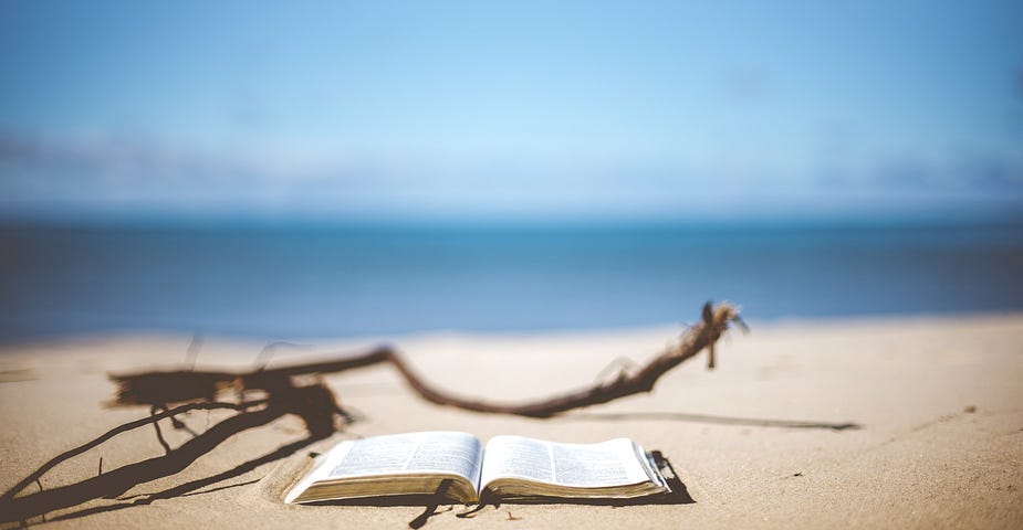 An open book lying on a sandy beach, with the water and sky visible in the background. No people are pictured nearby.