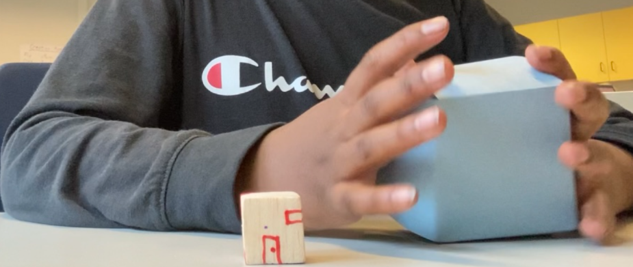 Hands of student folding 2D box while balsa wood house is in front.