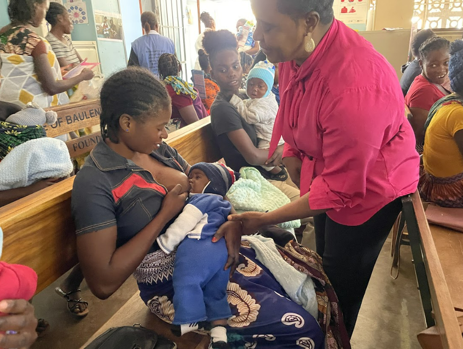 Nutritionist Esnart, wearing a bright pink blouse, gently helps a mother position her son to latch onto her breast, while another mother who is breastfeeding watches.