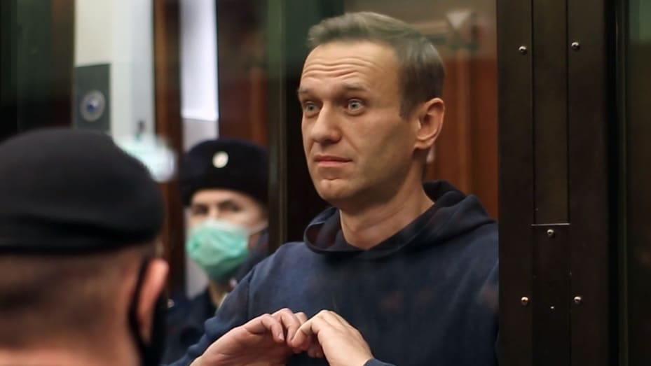 A photo of Alexei Navalny, a white man with blond hair wearing a blue hoodie. He is holding his hands in the shape of a heart on his chest behind glass, with a masked guard behind him.