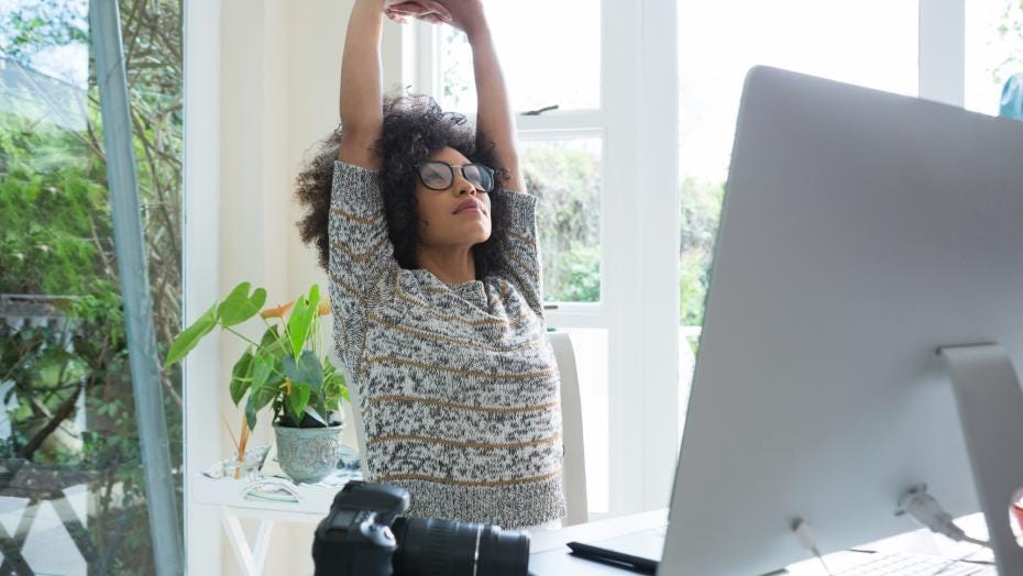 Woman with afro and glasses stretches her arms toward the ceiling in front of her work desk.