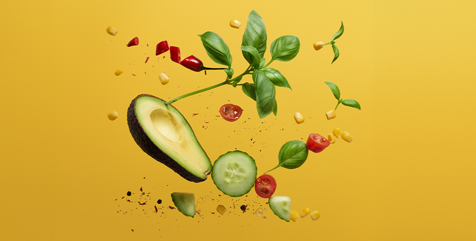 Avocado, cucumber, basil, and tomato looking as if they are being tossed over a yellow background while being sprinkled with spices