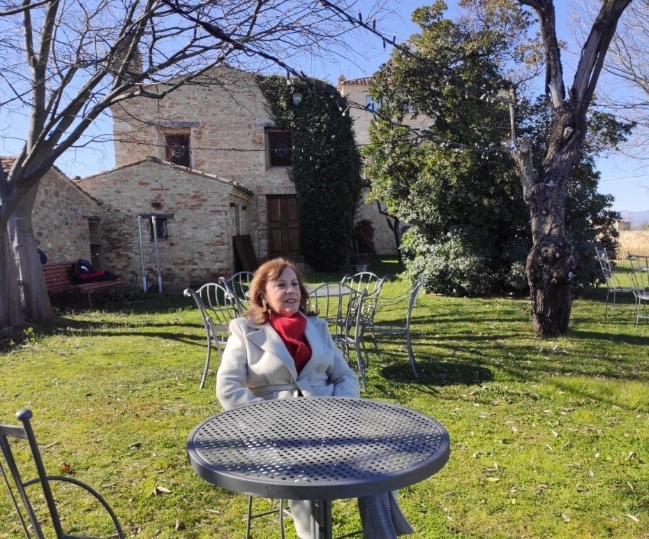 The author Judith Valente taking a moment to “do no thing” while seated at a metal table in a field with a stone farmhouse behind her.