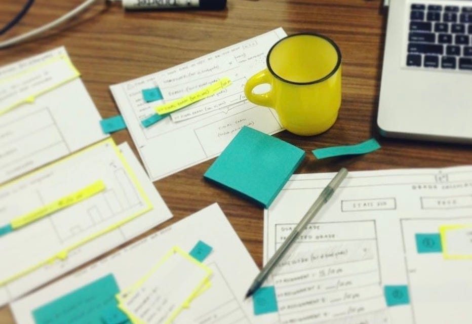 A desk covered in paper design prototypes, a yellow mug, blue post-it notes, a laptop, and a pen.