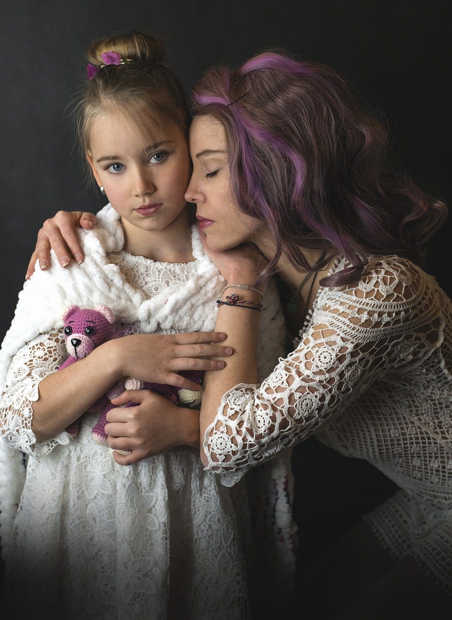 Mother and daughter, both dressed in white lace. The girl has small purple stuffed bear and a purple hair tie, the mother has purple highlights in her hair.