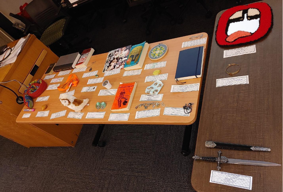 Image 1: Two rectangular tables with personal belongings placed on top, accompanied by hand written labels. Some of the belongings include a packet of Mentos, a stuffed toy snake, books, a cat collage, jewellery, a stuffed toy owl, and a dagger.