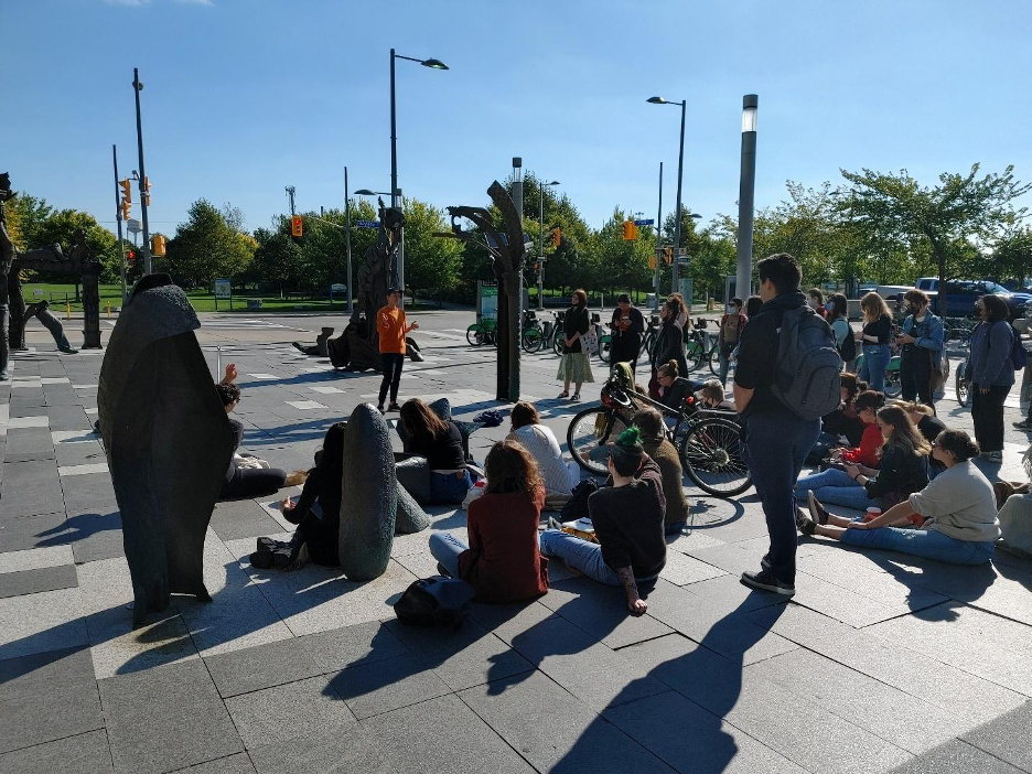 Image 2: On a sunny autumn day, a group of students sit and stand, listening to a white person speaking to them. The group is located at a public art sculpture that is cast from bronze, arising from a paved area at the edge of the crossroads of two streets in the Corktown Common area of Toronto, Canada. The sky is blue and the group cast long shadows that mix with the shadows from the sculptural pieces. In the background the trees and grass of Corktown Common are visible.