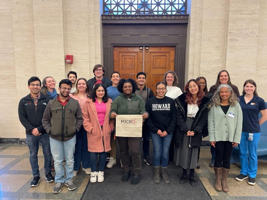 A group of people stand in front of a door. One person holds a banner that says “MICRO.”