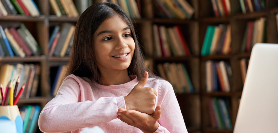 Young dark skinned girl with long dark brown hair, brown eyes and pink top signs “support”. Wall to wall bookshelves in background.