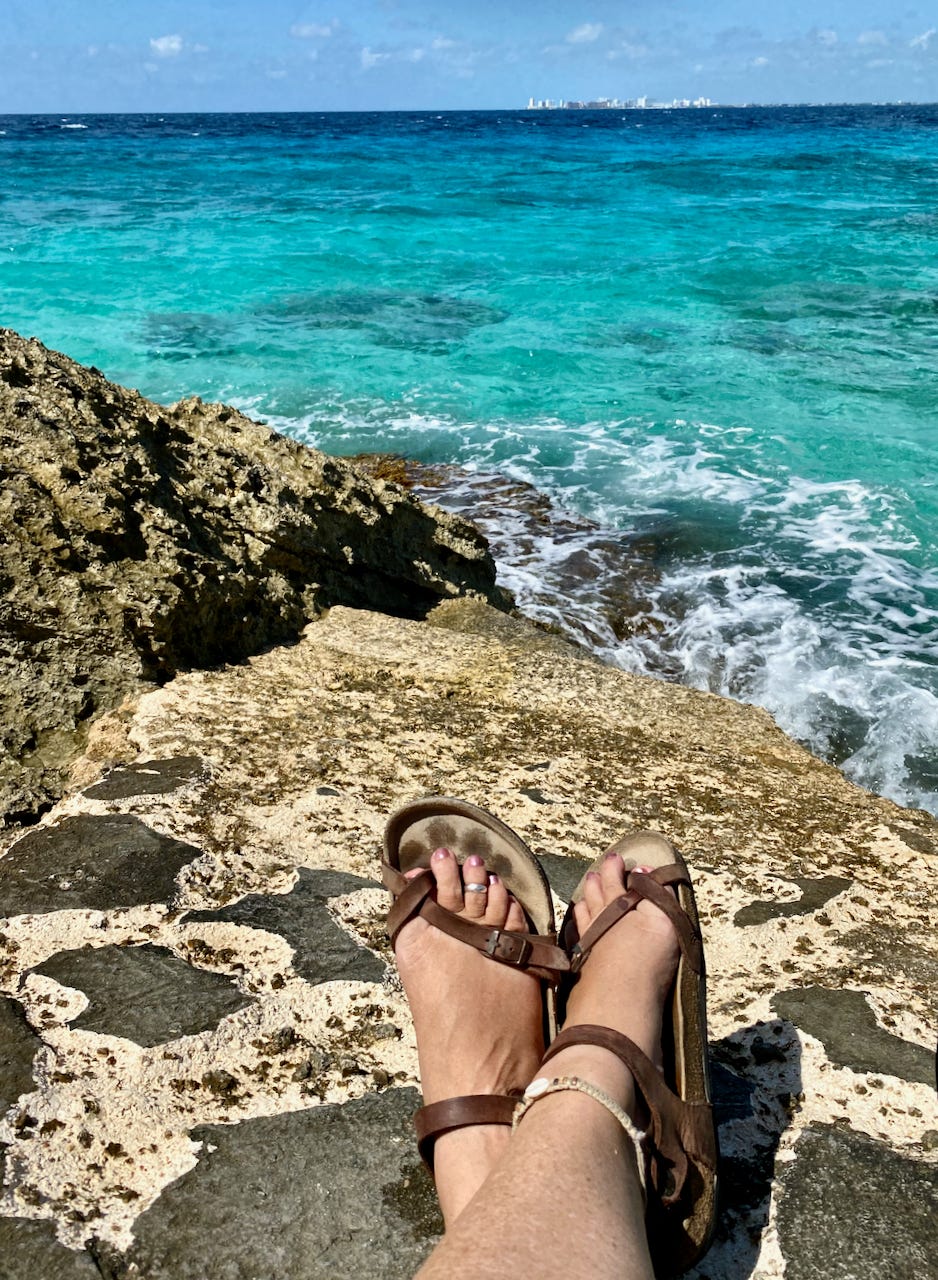 A lady's sandal-clad feet on a rocky shore where she sits looking out over the vibrant turquoise Caribbean water.