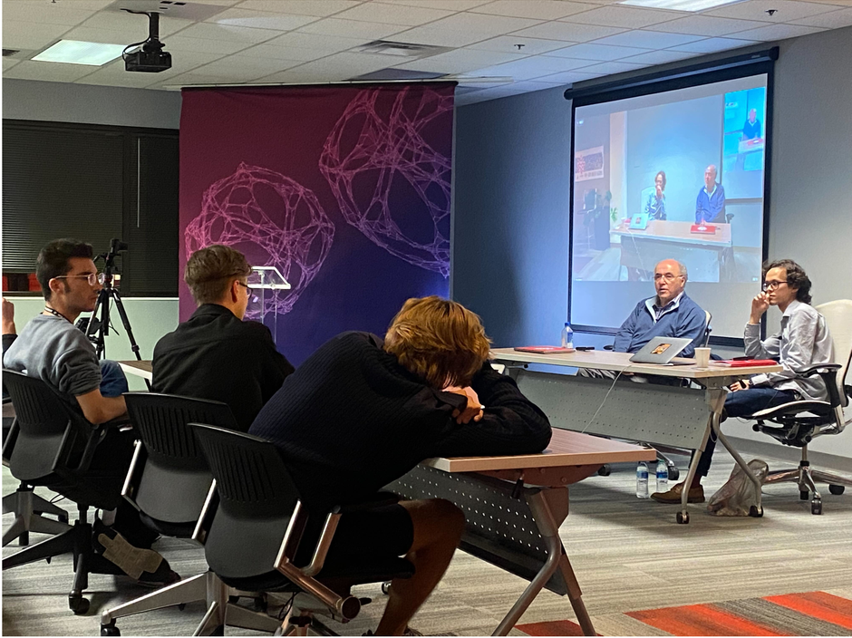 Students, mentors, and Stephen Wolfram meeting in a classroom to discuss things, with a protected video on a pull-out screen in the background