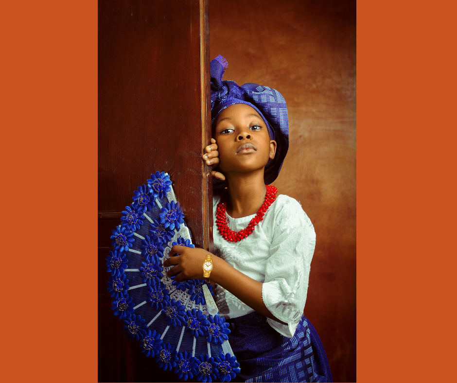 A young girl stands in an orange doorway, holding a blue fan and wearing a blue headdress.