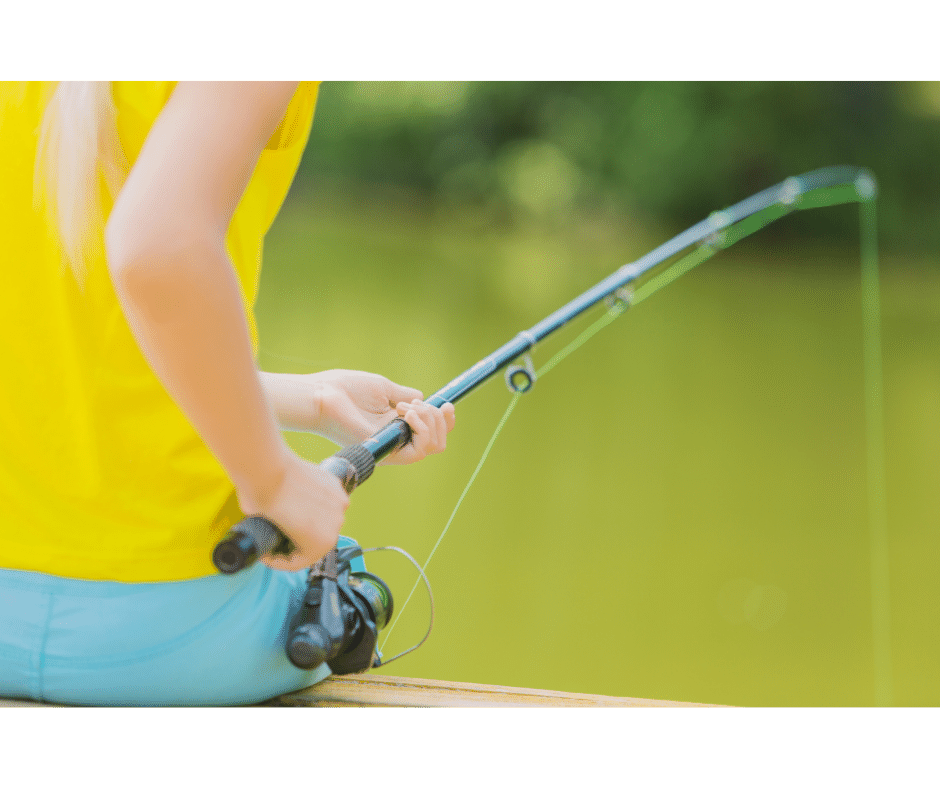 This image shows the body of a woman wearing a yellow sleeveless shirt sitting and holding a fishing rod cast on the water.