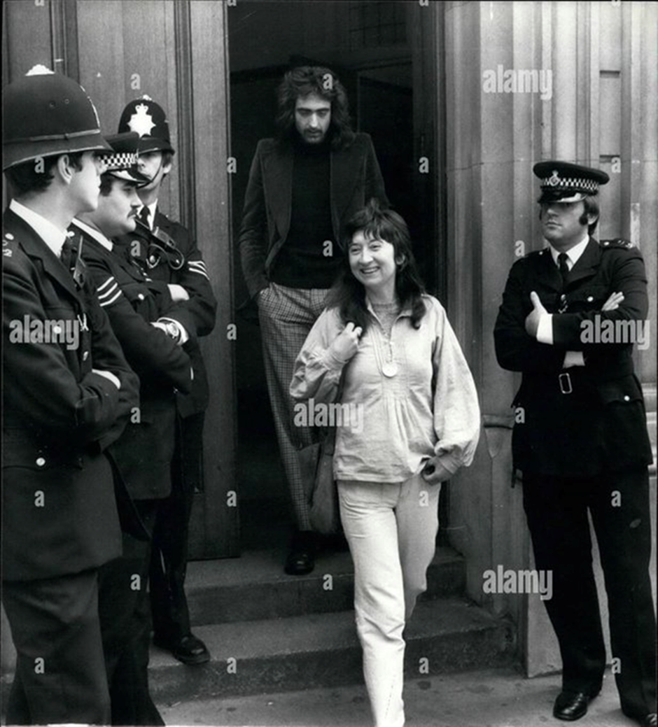 b/w photo shows a woman leaving a court building, smiling, flanked by police officers (not smiling)