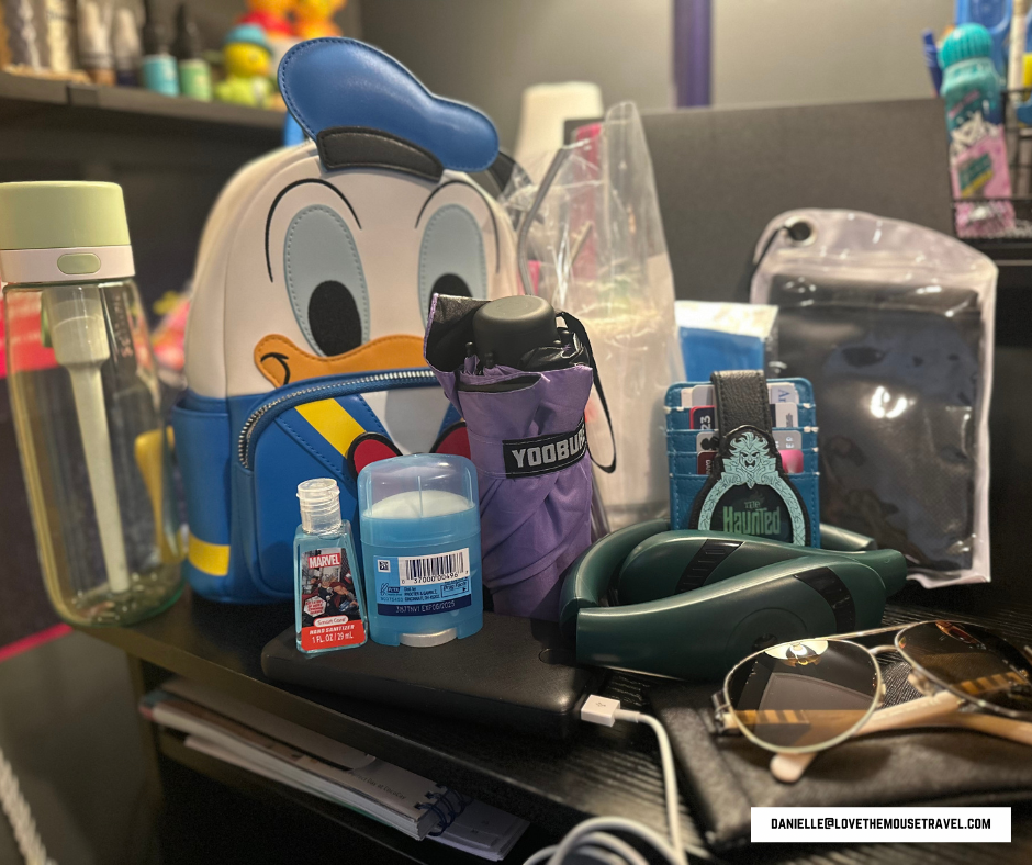 Image shows reusable water bottle, foldable umbrella, travel-size deodorant, travel-size hand sanitizer, portable battery charger, wallet, sunglass, and Donald Duck-shaped backpack for travel necessities Disney adult