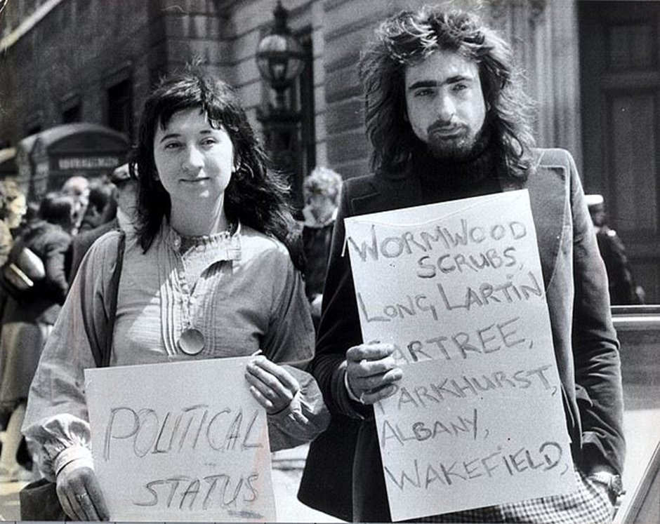 b/w 1970s shot of young couple in 70s clothes and hair holding up handwritten signs reading ‘political status’ and ‘Wormwood Scrubs, Long Lartin, Gartree, Parkhurst, Albany, Wakefield
