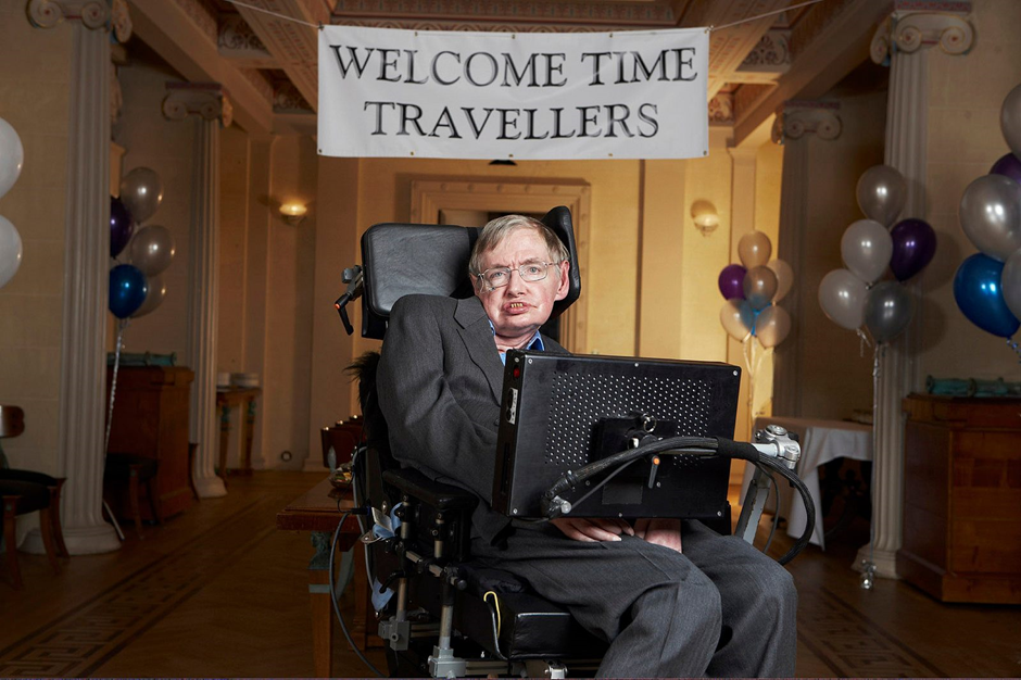 Stephen Hawking sits in his wheelchair in a posh university room under a banner reading “welcome time travellers”