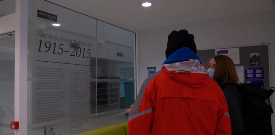 Screenshot from video, man in red and woman wearing a mask inside the University of Manchester Campus.