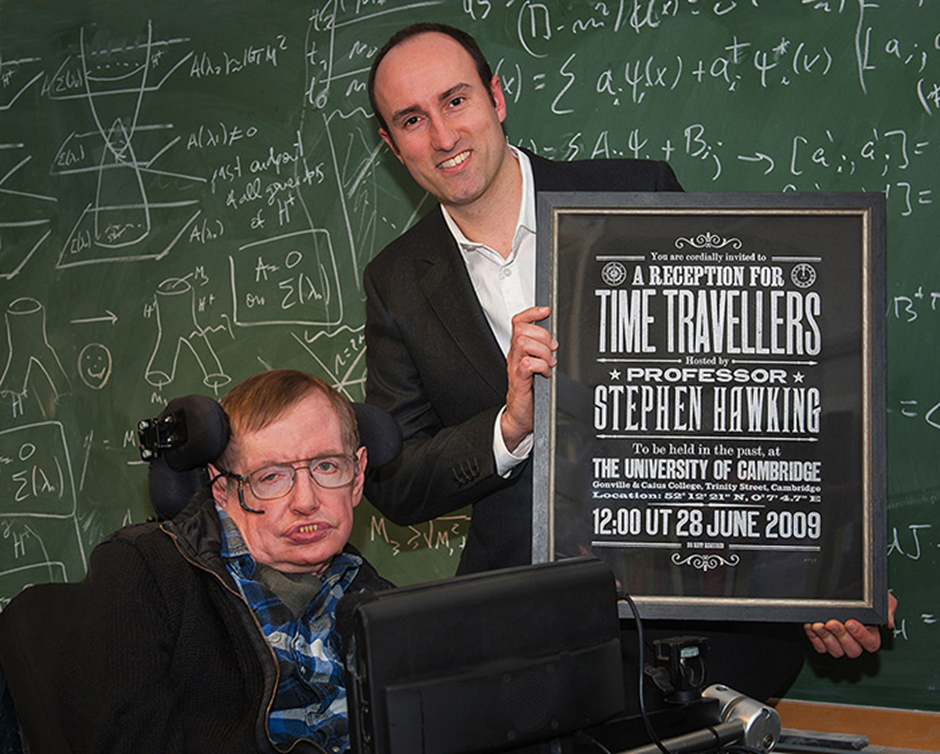 Stephen Hawking in his wheelchair, with a man standing next to him holding up a framed, hand-printed, limited edition of the time travellers reception invitation. It is designed and typeset to resemble a classic Victorian publicity poster