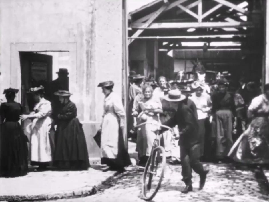 A group of workers leaving a factory building in 1895, one person with a bicycle.