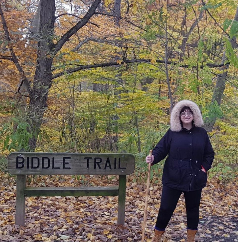 A white woman in her mid-twenties with glasses is standing next to a sign that reads “Biddle Trail.” Trees are visible in the background. The woman is dressed in black leggings and a black coat with a faux-fur hood. She is holding a walking stick and smiling.