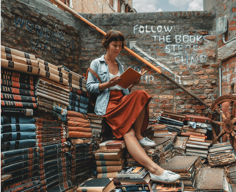 A woman reads a book will sitting on and around stacks of books.