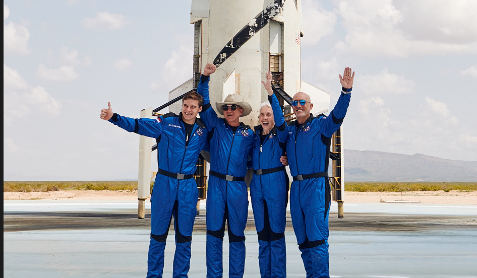 Jeff Bezos and his flight crew in front of the New Shephard rocket. Three men and a woman wave happily in blue space suits