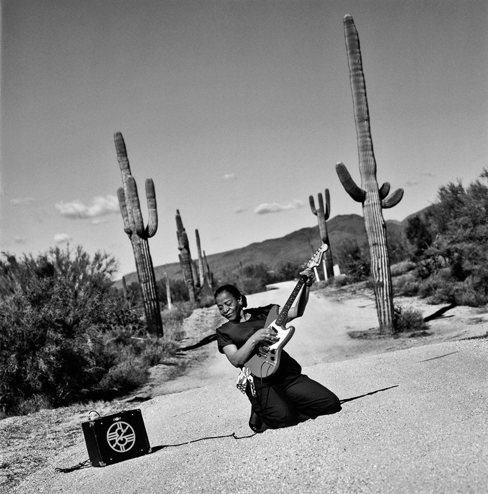 Beverly “Guitar” Watkins holding her guitar “Red Mama” in the middle of the dessert surrounded by cactuses and mountains.