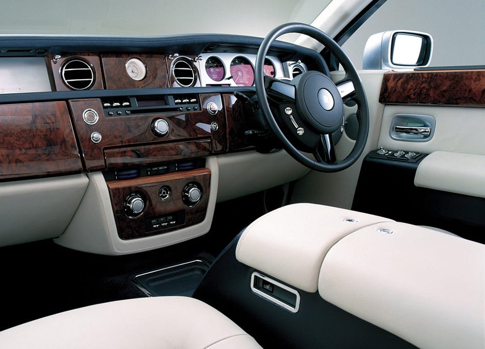 El interior de un Rolls-Royce Phantom de 2003, centrado en el lado del conductor. El tablero presenta paneles de madera y una variedad de controles y ventilaciones. Hay un volante en el centro y, a su derecha, una pantalla del sistema de infoentretenimiento rodeada de más acentos de madera. Los asientos están tapizados en cuero claro, y los paneles de las puertas a juego tienen manijas y controles integrados. La impresión general es de opulencia y alta calidad.