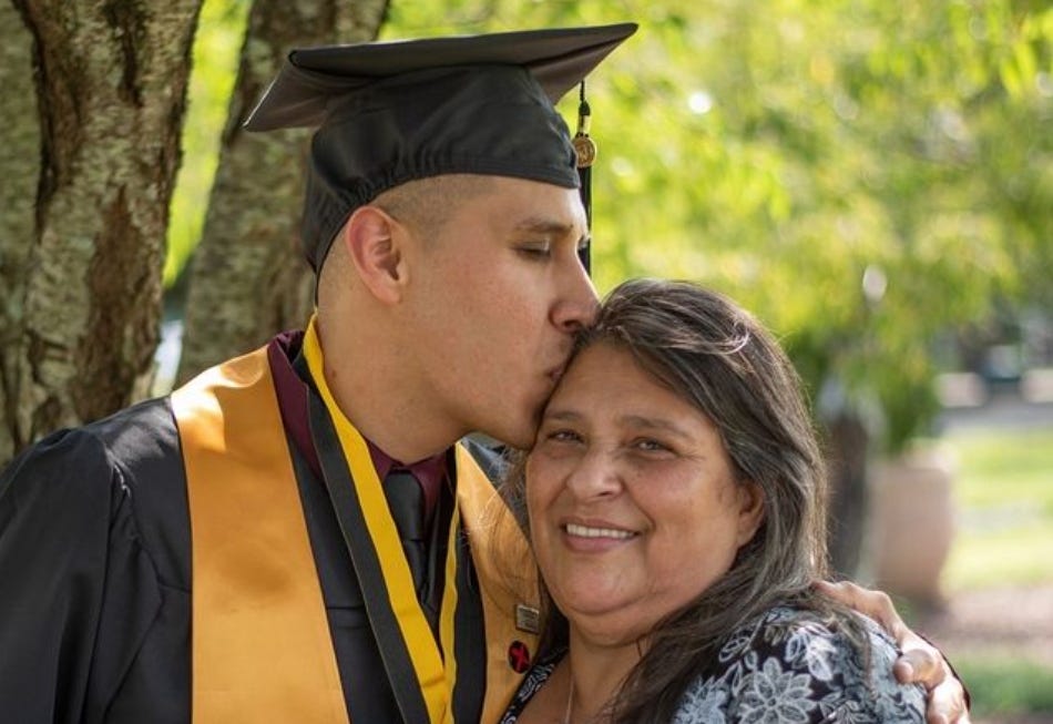 A graduate in a mortarboard kisses his smiling mother on the forehead.