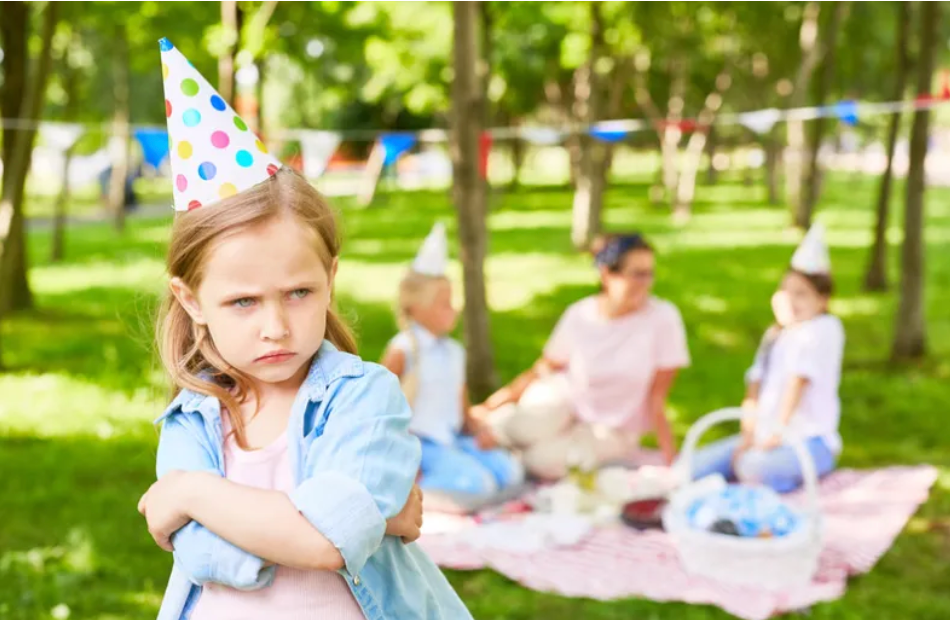 a little girl wearing a birthday hat, sulking. A mom and two other girls are sitting on a picnic blanket in the background.