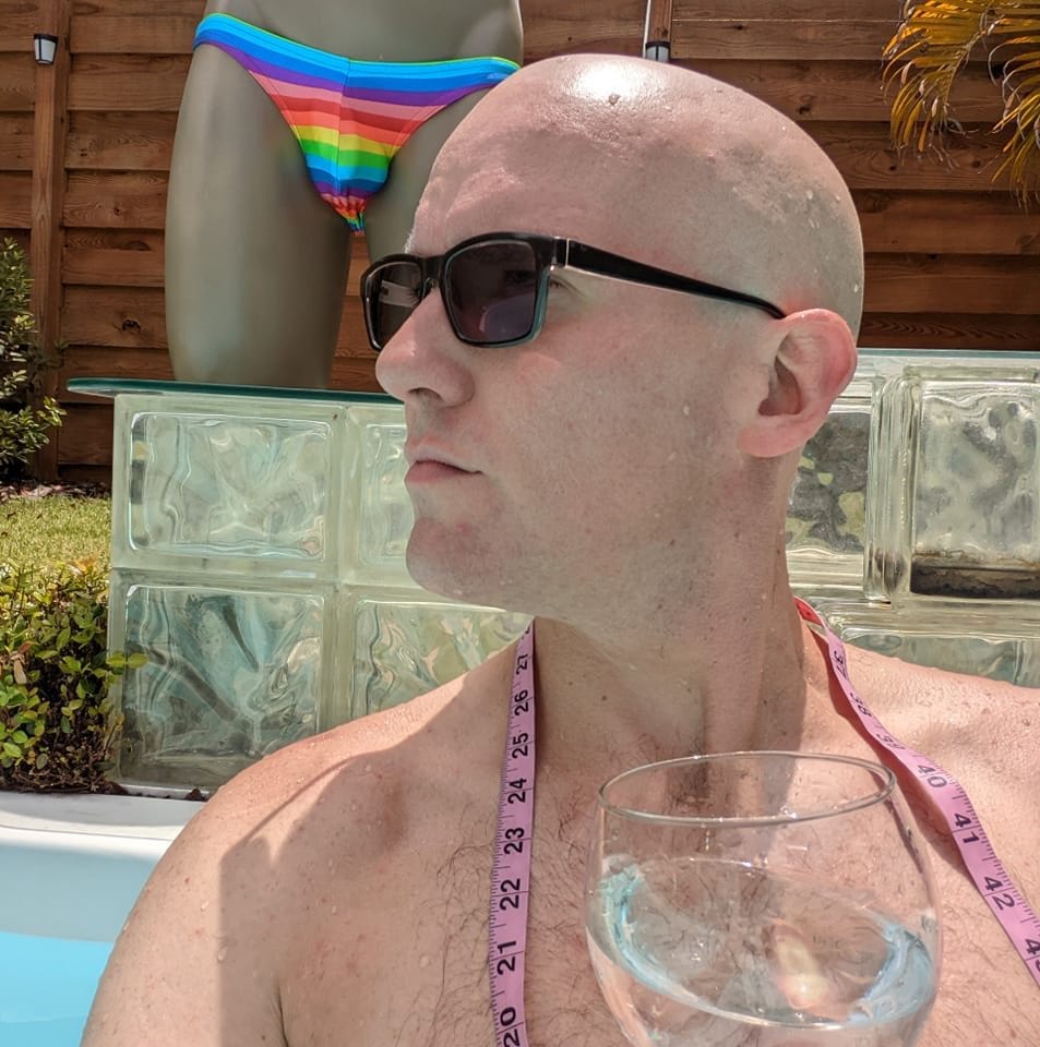 A shirtless, bald, white man wearing sunglasses is in a pool. He has a measuring tape draped over his neck and is holding a wine glass. In the background, there’s a mannequin wearing rainbow bikini bottoms.