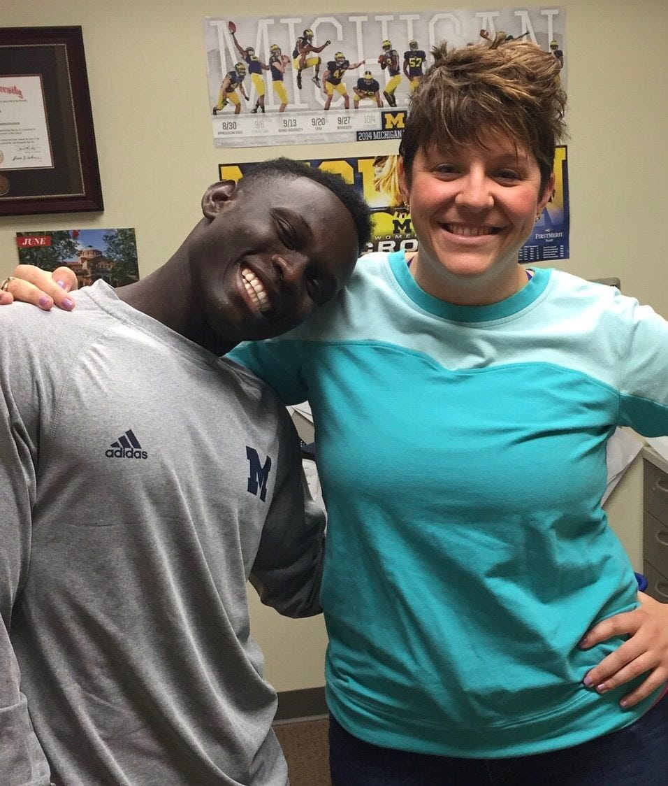 A man and woman side-hug and smile at the camera. A Michigan sports poster is behind them.