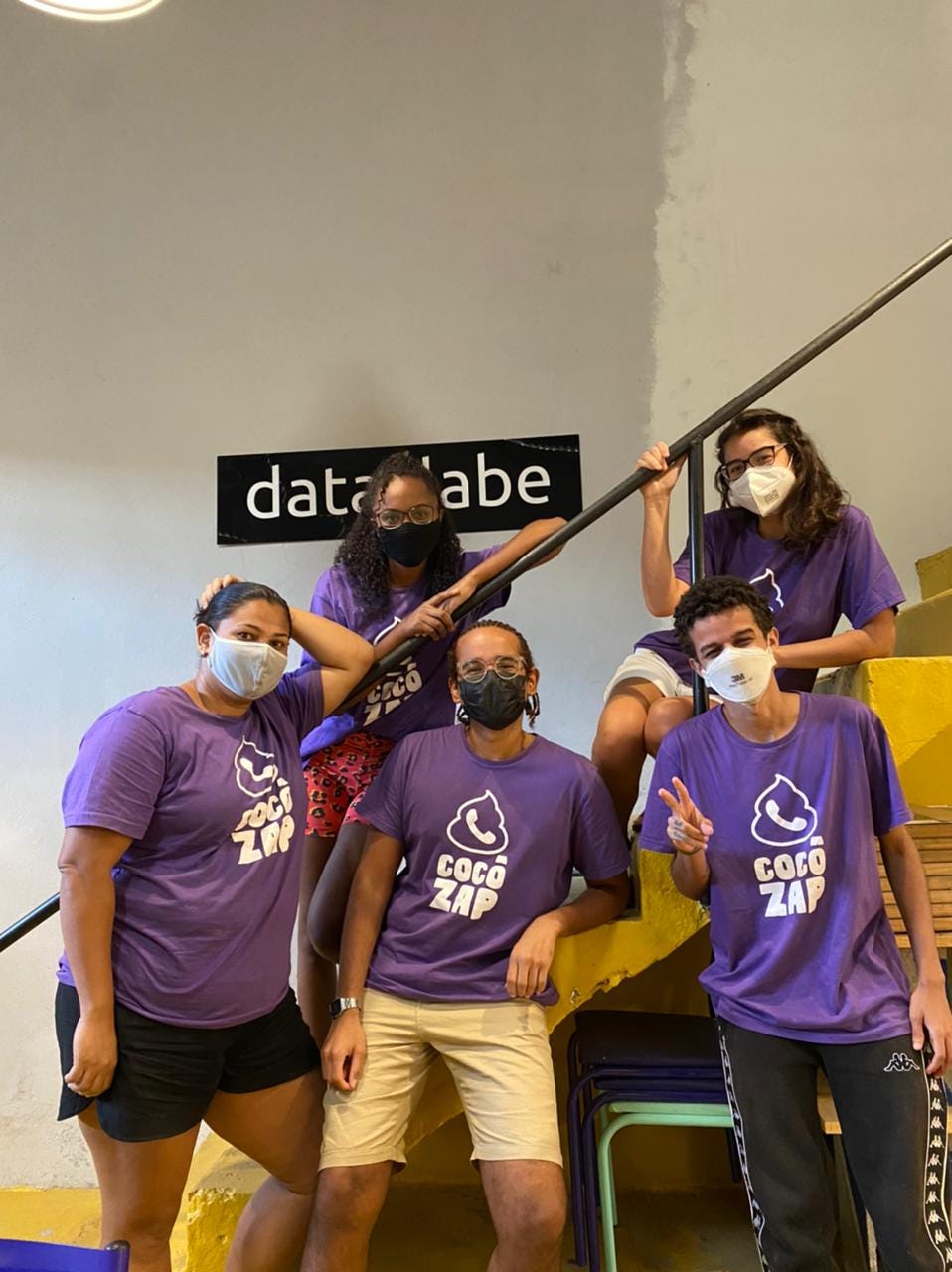 Embaixadores Cocôzap. Na foto estão 5 jovens, sendo 3 mulheres e 2 homens. Todos estão vestidos com uma camiseta roxa escrito "Cocôzap" e usam máscaras de proteção por conta da COVID-19. Estão posando em frente a uma escada amarela, e no fundo, há uma placa na parede escrito "data_labe".