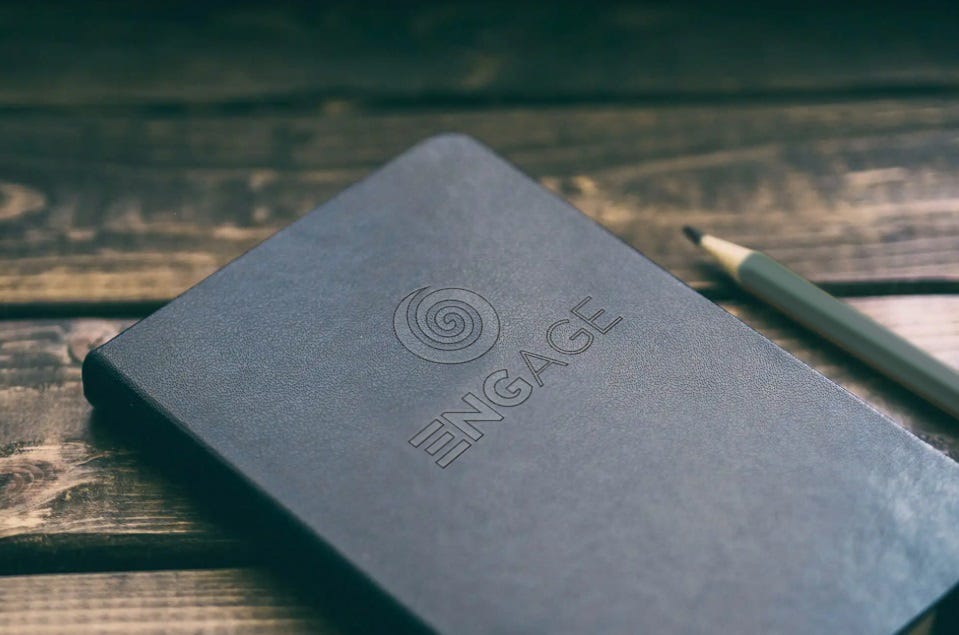 A black notebook with the Engage logo on the cover next to a pencil on a wooden table.