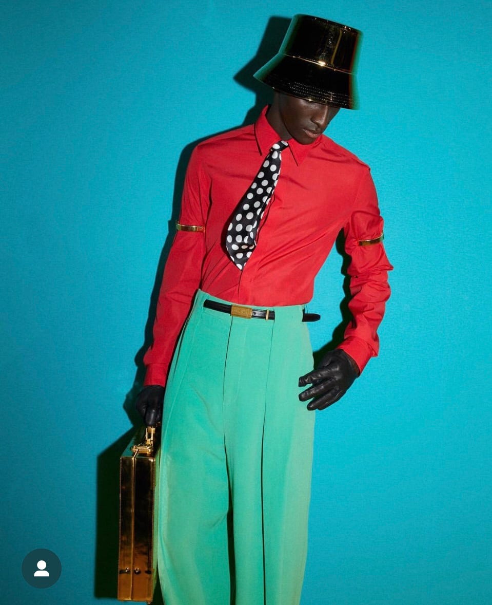 Balamain SS 2024. Man wearing a black patent leather bowl hat . A lon sleeve red shirt with a broad polkadot tie , leather gloves and a teal highwaisted trouser.