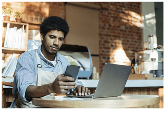 Black American male wearing an apron sitting inside a café with laptop and checking his cellphone while working