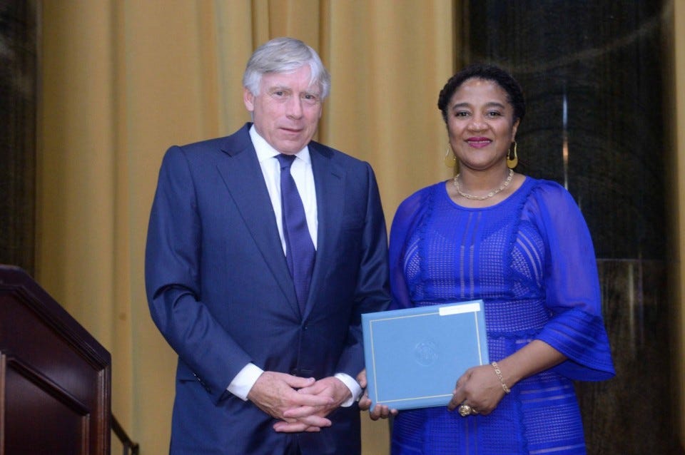 A man with gray hair and light skin in a blue suit stands with a woman with brown skin and hair in a blue dress. She smiles and holds a blue rectangular award — the Pulitzer Prize.