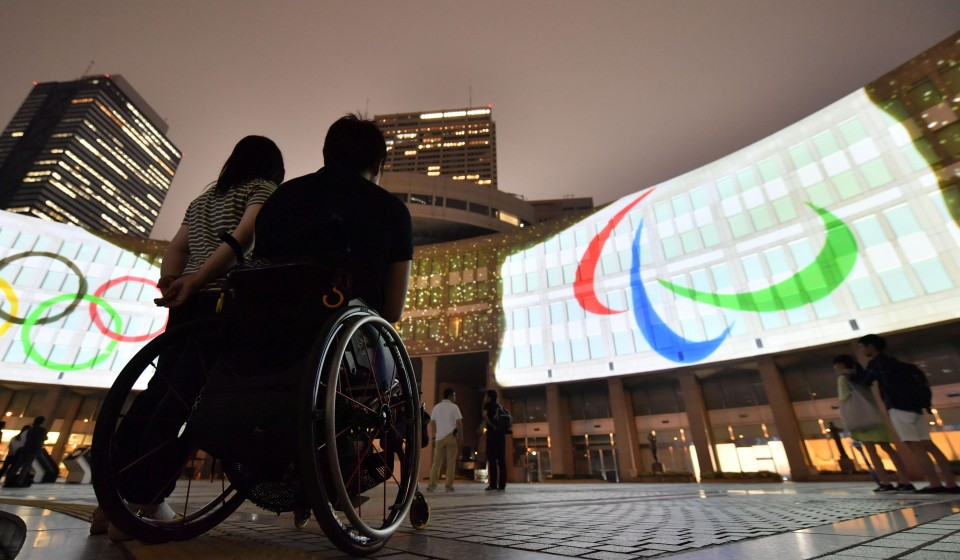 A Japanese man in a wheelchair gazing at the Paralympic sign displayed on a screen outside at night. Source: https://english.