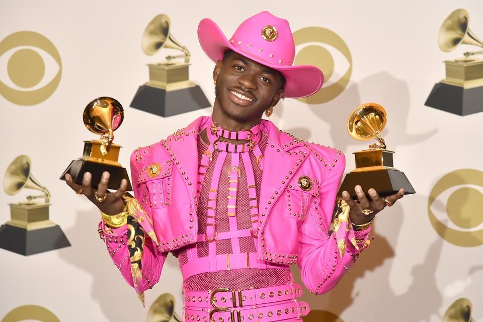 ID: Lil Nas X in a hot pink cowboy suit holding two Grammy awards