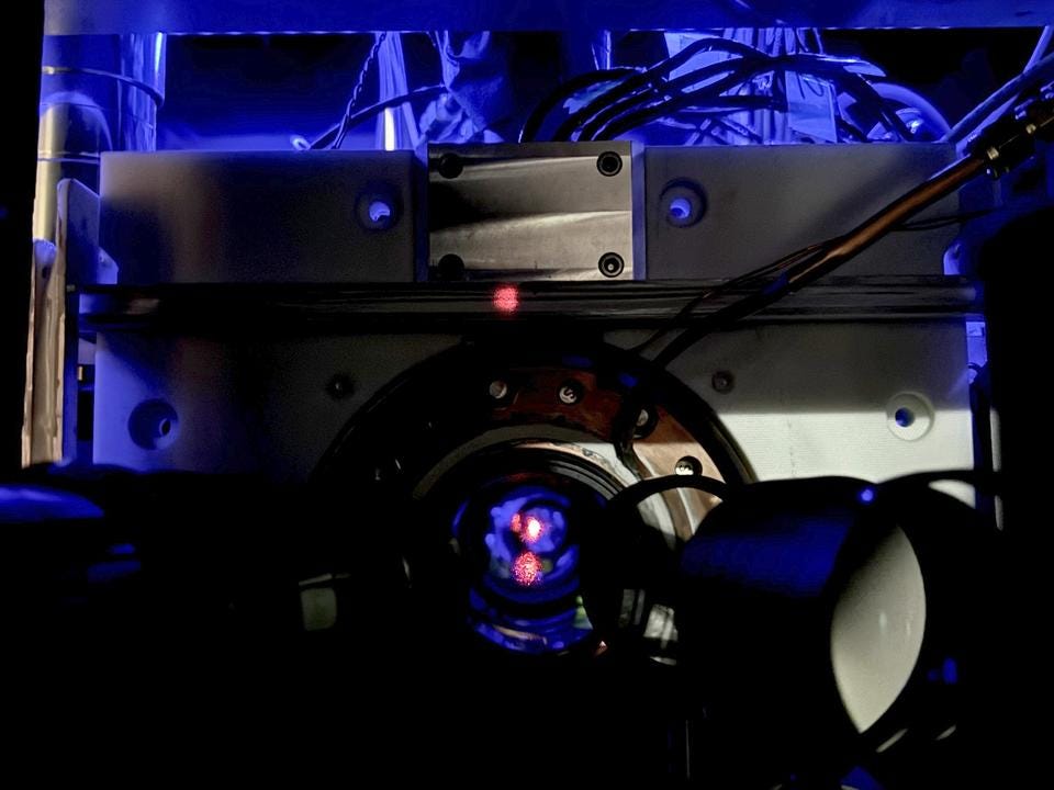 Close-up of a scientific instrument with wires and a circular metallic component, illuminated by blue light, featuring a small red dot at the center.
