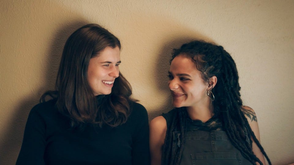 Two women in their twenties sit against a beige wall. One is a white woman with long dark hair, and the other is a light-skinned black woman with dreadlocks. They are both smiling.